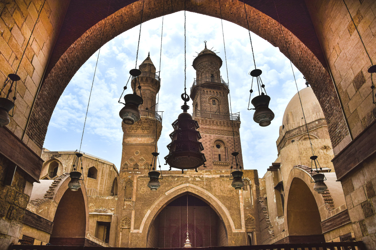 A view of two minarets and a dome on Moez Street through an arch with lamps hanging from it.