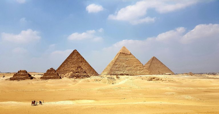 The pyramids of Giza stand golden in the setting sun against a a blue sky.