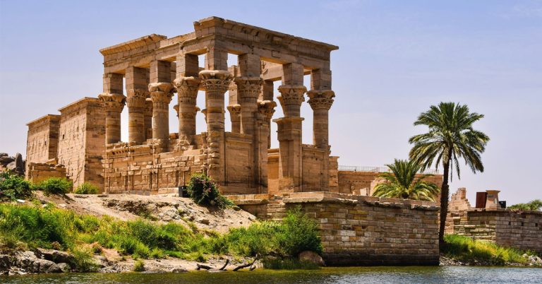 The Philae Temple in Aswan is shown with the rows of columns surrounded by greenery and the Nile waters in the foreground.